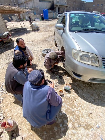 This image depicts an outdoor scene in کوکران, Afghanistan, featuring five men around a silver four-door sedan on an unpaved, rocky, and dusty surface. A man dressed in a light brown outfit and cap is actively squatting and working on the front passenger side wheel assembly of the vehicle, which has its tire and wheel detached and resting behind him on the ground. Three other men are seated on the ground to the left of the mechanic. One man, wearing a checkered shirt and dark vest, observes the repair. Another man, in a dark outfit and vest, holds a dark-colored smartphone. A third man, wearing a blue outfit, vest, and a blue patterned skullcap, is observed looking down at several objects on the ground, including a small open container of a viscous substance, a small cardboard box, and coiled wires. In the immediate foreground, a red and white plastic jug lies on its side. In the background, simple, dilapidated buildings constructed from concrete blocks are visible under a bright sky, some with rudimentary awnings or thatched roofs. A large blue plastic water tank stands near one of the buildings. A motorcycle is partially visible on the far left. A distant figure stands near a background structure. The scene is illuminated by direct sunlight, casting distinct shadows.