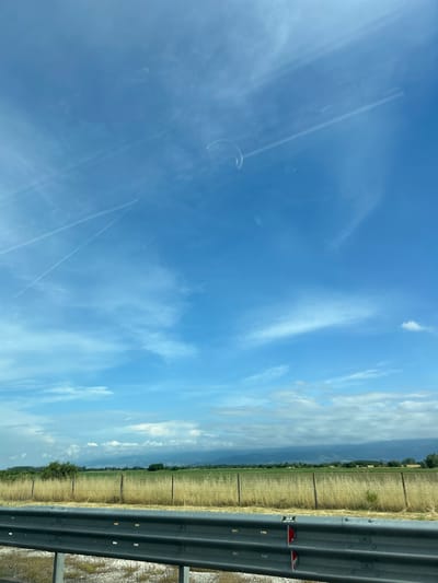 This media file captures a sunny daytime landscape from the perspective of a moving vehicle, likely in Malo Konare, Bulgaria. The image, originally rotated, shows a wide-angle view dominated by a clear blue sky with scattered white, wispy clouds and several faint airplane contrails. Prominent glare and reflections on what appears to be a car window are visible across the sky, including faint circular and linear patterns.
In the right foreground, a dark grey, corrugated metal guardrail runs horizontally across the frame, indicating a road or highway. Attached to the guardrail is a small white rectangular marker with black text, partially visible as "T 200" and possibly "196" or "1967" below it. Below this, two red reflective strips are also affixed to the guardrail.
Beyond the guardrail, a dry, golden-brown field stretches into the midground, traversed by a line of fence posts. In the distance, a hazy horizon line reveals a band of green trees and distant hills or mountains. The weather appears bright and clear, characteristic of a sunny day, possibly during the summer or a dry season given the color of the fields. The scene conveys a sense of travel through a rural, open landscape. No individuals are visible in the image.