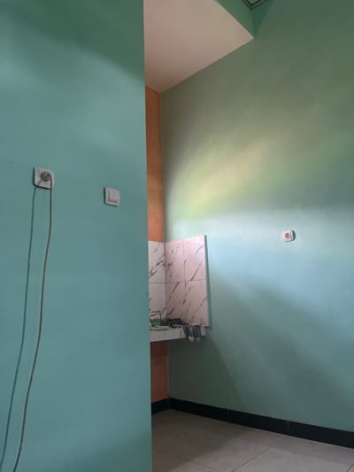The image depicts an indoor scene with walls painted in various shades of turquoise and a section of peach-colored wall. In the foreground, a turquoise wall occupies the left side of the frame, featuring a white electrical outlet with a grey wire plugged into it, extending downwards. To the right of this outlet is a white light switch.

The background reveals a corner where the turquoise wall meets a peach-colored wall, which then transitions to a larger section of the turquoise wall. A tiled backsplash, with a marble-like pattern of white and grey squares, is visible above a countertop. On the countertop, several items are present, including what appears to be a small box or package with the word "mama" visible, and a pair of grey socks hanging over the edge.

Further to the right, on the larger turquoise wall, another white electrical outlet is present. Sunlight is casting shadows and creating a gradient of light on this wall, transitioning from a brighter yellow-green near the ceiling to a lighter turquoise towards the floor. The floor itself is tiled with large square tiles in a neutral tone.

The scene appears to be a domestic interior, possibly a kitchen or utility area, located in Meteseh, Indonesia, given the context. There are no people visible in the image.