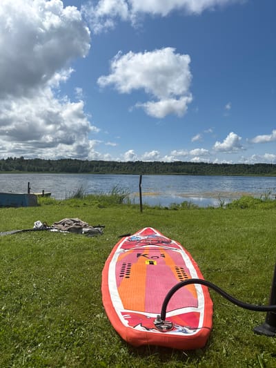 In Chikino, Russia, on a bright, sunny day, a red and orange stand-up paddleboard (SUP) is laid out on a grassy shore, connected to a black air pump by a flexible hose. The SUP is positioned in the foreground, facing away from the viewer. To the left of the SUP, a rolled-up blanket or towel and a paddle are visible on the grass. Beyond the grassy area, a calm lake stretches out, bordered by a dense forest in the distance. The sky above is a clear blue, dotted with fluffy white clouds. A small blue boat and a wooden pier are situated near the water's edge. The overall scene depicts preparations for a watersport activity in a serene, natural setting.