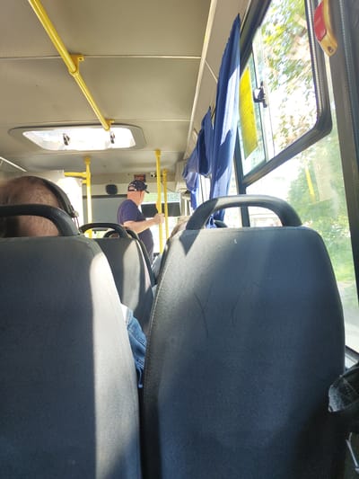 Inside view of a bus interior, looking towards the front. Several dark grey seats with black handles are visible in the foreground. Sunlight streams in from the right side window, illuminating the seats and the aisle. In the middle ground, a man wearing a dark blue t-shirt and a blue baseball cap stands near the front of the bus, holding onto a yellow vertical pole. He appears to be looking forward or to his left. Blue curtains hang near the window on the right. The ceiling of the bus features yellow horizontal and vertical poles. Outside the window, green trees and a building can be seen. The image is taken from a passenger's perspective, seated in the back of the bus.