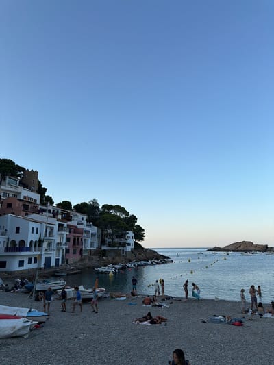The image depicts a coastal town in Begur, Spain, characterized by its picturesque Mediterranean architecture. White and pastel-colored buildings, adorned with balconies and arches, cascade down a hillside towards a pebble beach and a calm, blue bay. Several small boats are moored in the water, and a few larger yachts are visible in the distance. The beach is populated with people, some sunbathing on towels, others wading in the shallow water, and a small group engaged in a casual game on the shore. Lush green trees dot the landscape, providing a natural contrast to the built environment. The sky is a clear, pale blue, indicating a pleasant, possibly late afternoon or early evening atmosphere. In the distance, a rocky island rises from the sea, adding to the scenic beauty of the location. A medieval tower stands prominently on the hillside, hinting at the historical significance of the area.