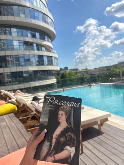 A person holds a book titled "Roxolana" by Pavlo Zagrebelnyi in front of a swimming pool and a modern building on a sunny day. The building, with its curved glass facade, appears to be a hotel or residential complex. The pool area is equipped with lounge chairs, some occupied by people relaxing. The sky is blue with scattered clouds, and lush green trees are visible in the background. The overall scene suggests a luxurious urban setting, possibly in Kâğıthane, Turkey, given the building's architecture which is characteristic of modern developments in the area.
