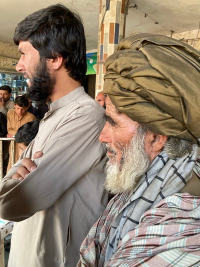 The image shows two men indoors, presumably in Qilla Saifullah, Pakistan. An older man is partially visible, wearing a large, olive-colored turban. His face shows age, with a white beard. He also wears a multi-colored, patterned cloth, draped on top of the turban. The second man is younger, with dark hair and a beard. He wears a light grey, collared shirt. Multiple other people are visible in the background, though they are partially obscured. The environment appears to be a shop. Shelves filled with merchandise are visible behind the men.