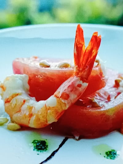 A close-up, slightly out-of-focus shot shows a dish of food served on a white plate. The dish consists of a cooked shrimp, its tail section curled around two slices of red tomato. The tomato slices are topped with what appear to be pine nuts. A dark sauce line and a small dollop of green sauce are visible on the plate. The background is blurred, showing patches of green and light blue, possibly indicating an outdoor setting with foliage and sky. There is no indication of a specific location in Vitoria-Gasteiz, Spain, or any people present.