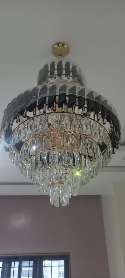 The image shows a decorative crystal and black metal chandelier mounted on a white wall. The chandelier has a spherical shape with multiple layers of rectangular crystal prisms and black metal accents. It is attached to the wall by a circular golden mount. The chandelier appears to be new, with some plastic protective wrapping still visible on its outer layers. The setting appears to be indoors, possibly a room in a house or a commercial establishment. The lighting suggests it is daytime, and there are no obvious weather cues. The background includes a light purple wall and a dark-framed window with a view of the interior of a building. There is no visible text in the image. The overall impression is one of luxury and modern design.