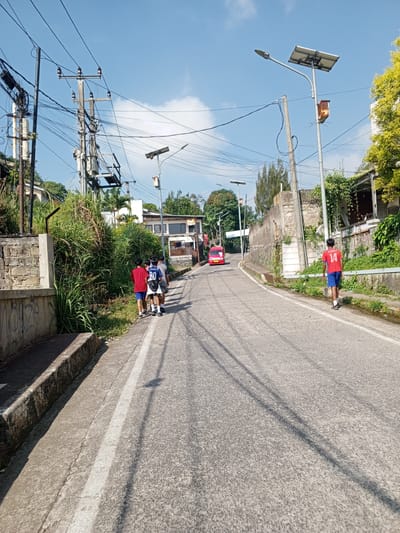 This image depicts a residential street scene in Weddagala, Sri Lanka, on a clear, sunny day. The paved, single-lane road, composed of light grey asphalt or concrete, ascends gradually from the foreground into the distance. A faded white line demarcates the left edge of the traversable road.
On the left side of the road, a concrete retaining wall with some visible graffiti borders an elevated area densely covered with tall, green vegetation. Beyond the vegetation, multiple utility poles, laden with a complex network of overhead electrical and communication wires, line the street. One prominent pole features a transformer unit. A group of four young males is observed walking away from the camera on the left shoulder of the road. One wears a blue shirt with "ADIYA 20" on the back.
On the right side of the road, another concrete wall, partially covered with climbing vines, defines the street edge. Beyond this wall, green vegetation and parts of residential structures are visible. Several streetlights equipped with solar panels are installed along this side, with one prominent streetlight featuring an orange-brown box mounted below its solar panel. A single young male, wearing a red shirt with the number "14" on the back and blue shorts, walks away from the camera on the right shoulder of the road.
Further up the road, a small red mini-van, possibly a public transport vehicle, is visible traveling in the same direction as the pedestrians. The sky is largely clear blue with scattered white cumulus clouds. Bright sunlight casts distinct, elongated shadows of the utility poles and wires across the road surface, indicating strong illumination.