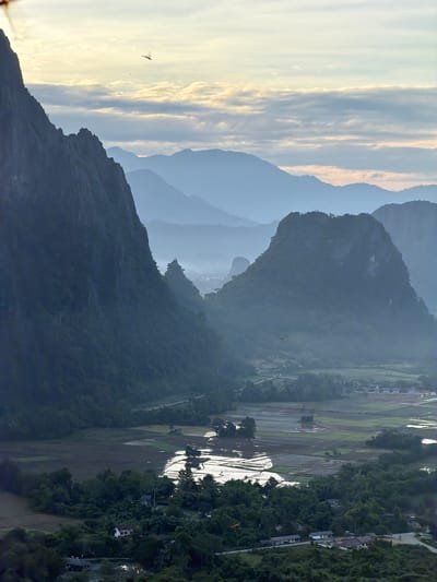 This is a vertical, eye-level, outdoor, wide-angle shot of a misty, mountainous landscape at sunrise. The foreground is dominated by a steep, dark green, heavily vegetated mountain range on the left, casting a shadow over the valley below. In the valley, there are patches of cultivated fields, some reflecting the soft, hazy sunlight and appearing as pools of water. Scattered throughout the valley are a few small houses and buildings, partially obscured by trees. In the mid-ground, more rounded, smaller mountains rise, also covered in dark green vegetation and shrouded in mist, creating a layered effect. In the background, distant mountain ranges fade into a pale blue haze under a soft, pale yellow and white sky with thin, horizontal clouds. A small, red dragonfly is captured in flight near the top left of the frame, and another smaller, indistinct object, possibly another insect, is also visible in the sky. The overall atmosphere is serene and slightly mysterious due to the mist and soft lighting. The location is Naka, Laos, a region known for its karsts and rice paddies.