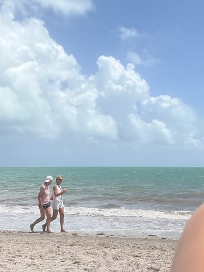 The image depicts two individuals on a beach. The environment includes sand, ocean water, and a sky with clouds. The sky takes up most of the frame with bright blue interspersed among white puffy clouds. The sand and surf run lengthwise with two people in the middle of the frame. One individual is wearing a pink shirt and dark-colored shorts with a white hat. The other individual is wearing a white top and white shorts. It appears the location is a beach in Key Biscayne, Florida, based on the landscape and climate.