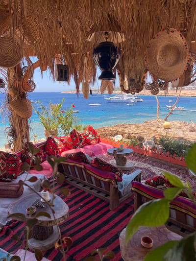 This image depicts an outdoor seating area with a view of the Red Sea, likely in Sharm El Sheikh, Egypt. The seating area is covered by a thatched roof, adorned with various woven baskets, hats, and decorative lamps. Below the roof, comfortable seating is arranged with numerous colorful cushions featuring intricate geometric patterns. Red and black striped rugs cover the floor, and a larger patterned rug is visible beneath the seating.
In the background, the crystal-clear turquoise waters of the Red Sea are visible, dotted with several boats and yachts. The coastline features buildings and lush greenery. The overall atmosphere suggests a relaxed, bohemian-style setting, possibly a cafe or lounge area overlooking the sea. The natural light and vibrant colors contribute to a sense of tranquility and vacation.