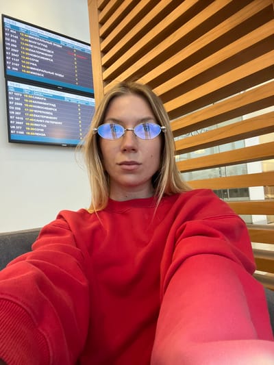A person with blonde hair and reflective glasses is shown in a selfie, wearing a red sweater. Behind the person, there is a wooden slat wall. A flight departure board is visible with Russian text indicating flight details, times, and destinations. Destinations listed include cities like Volgograd, Mineralnye Vody, and Ekaterinburg.