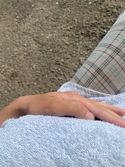 A close-up shot captures a person's hand resting on a light blue towel. The hand, with visible fingernails, lies horizontally across the textured fabric. Behind the towel and hand, a person's legs are visible, clad in plaid pants. The background consists of a grainy, brown, gravelly surface. This image appears to be taken in Myrza-Ake, Kyrgyzstan, given the context.