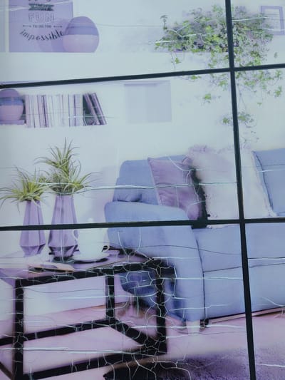 This is an indoor photograph, likely taken in a living room or display area, possibly in Bielany Wrocławskie, Poland. The scene features a light blue sofa with several cushions, some appearing to be decorative and textured with fringe. A glass-topped coffee table sits in front of the sofa, holding two modern, faceted purple vases with green, spiky plants, and a white teapot on a saucer.
On a shelf above and behind the sofa, a row of books is visible, along with decorative purple spheres. Further up and to the left, a framed poster with pastel colors displays text. The words "Kind of FUN" are prominent, with "TO DO THE" below it, and "impossible" in a stylized script beneath that.
The background wall appears to be a light, neutral color. To the right of the sofa, a hanging plant with green leaves cascades downwards, casting subtle shadows. The overall color palette is soft and pastel, dominated by light blues, purples, and greens, creating a serene and stylish atmosphere. The image is overlaid with a distinct pattern of cracks, suggesting the photograph might be on a damaged surface or a deliberate artistic effect. No people are visible, and the lighting suggests an indoor setting, possibly during the daytime.