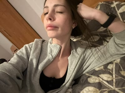 A woman wearing a gray zip-up hoodie over a black top is lying on a patterned blanket. She has long, dark hair and is wearing a black smartwatch on her left wrist. Her head is tilted, and she is winking with her right eye, lips slightly parted. A wooden dresser or cabinet is partially visible in the background.