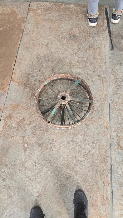 The image, taken in Gangapur, India, presents an overhead view of two people standing on a textured ground surface, with a heavily weathered circular object positioned between them.
The ground is a flat expanse of what appears to be concrete or a large metal plate, showing a mottled grey-brown coloration and faint linear seams. To the left, the lower legs and feet of one person are visible, wearing light-colored pants and black and white shoes with prominent white soles. To the right, the feet of a second person are visible, clad in dark, possibly black, robust shoes. Both individuals are positioned around the central object.
The central object is a circular, aged structure, strongly resembling an old wheel or a turbine component. It features a central hub with a hole, from which numerous spokes or blades radiate outwards to a circular rim. The object shows significant signs of corrosion and rust, displaying reddish-brown tones. Some of the spokes have remnants of a greenish-blue material or paint, indicating previous color.
The lighting is bright and even, suggesting it is daytime. No specific activity beyond standing is discernible, and there are no emotions or visible text captured in the frame. The scene conveys an impression of an aged, possibly industrial or rural outdoor environment.