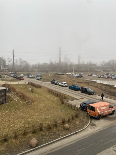 This image depicts a foggy, overcast daytime scene in Новогорелово, Russia, with an industrial background and an urban/suburban foreground.
In the far background, obscured by heavy atmospheric haze, stand multiple tall, brick-colored industrial chimneys, at least three of which are clearly visible, with associated industrial structures. Plumes, possibly steam or smoke, are emitted from at least one chimney. Utility poles and power lines traverse the mid-distance.
The midground features a network of paved asphalt roads and unpaved dirt/muddy areas functioning as parking lots. Numerous vehicles are present; a queue of cars (a black SUV with a roof rack, a white SUV, a blue sedan, and another black SUV) is visible on the paved road in the lower-mid right. An orange commercial van with the word "IEDER" partially visible on its side is parked on the curb in the lower right, behind a black passenger vehicle. Many other cars of various makes, models, and colors (including red, white, grey, and black) are parked both on paved sections and scattered across the unpaved, muddy ground. A small parking sign with a white 'P' on a blue background is visible near the center-left.
The ground cover is predominantly dormant, light brown grass, interspersed with patches of bare earth and mud. Sparse, leafless deciduous trees and low, dormant shrubs line the roads and unpaved areas. A paved pedestrian pathway, bordered by low shrubs and containing at least four streetlights, runs parallel to the main road in the foreground-left. Two large, light-colored boulders are positioned as landscape features in the lower-mid left. Part of a building, featuring a patterned stone patio, is visible in the extreme lower-left corner.
One individual, dressed in dark clothing, walks away from the camera on the right side of the paved road, carrying a white bag. The sky is uniformly light grey, indicating heavy cloud cover and haze, resulting in diffused lighting.