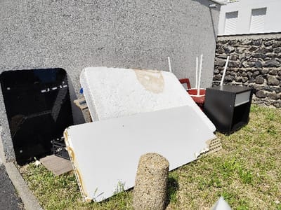 The image shows discarded furniture and other items in front of a grey concrete wall, likely an apartment building. The wall is located in Clermont-Ferrand, France, at coordinates (45.787, 3.11). The items include a white mattress, a large white rectangular board, a black rectangular glass object, a dark brown wooden nightstand, and a partially visible red bucket. The items are lying on a grassy area. The nightstand has a white drawer. The white board has a slightly chipped corner. There is a rough, medium-sized grey stone visible at the bottom of the image. The nightstand is positioned against a stone wall. There is a partially visible white wall with a roller shutter window on the right side of the image. The items appear to have been left there by the residents of the building, possibly as part of a cleanup effort or waste disposal.