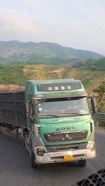 A green and silver HOWO truck is parked on a road. The truck's license plate reads "47E 007.67". In the background, a landscape of hazy green mountains is visible. The scene appears to be located in a rural or mountainous area, with evidence of terraced agriculture on the hillsides. While the image shows a truck, mountains, and road, specific details confirming the location as Aê Thi, Vietnam, are absent.