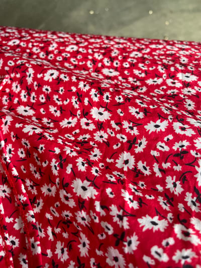 The image shows a close-up of a red fabric with a small white and black floral print. The fabric is creased and appears to be draped over a surface, creating folds and shadows. The background is out of focus and appears to be a neutral, textured surface, possibly a wall or a table. There are no people, animals, or identifiable landmarks visible in the image. The location in the city of Zanzi, Nigeria is not discernible from the visual information provided.