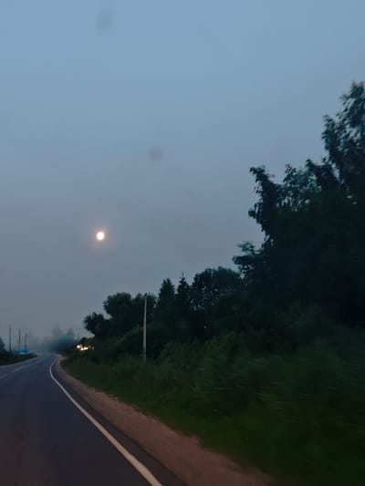 The image depicts a road in Batiushkovo, Russia, during twilight or at night. A full moon is visible in the hazy sky. The road curves to the left, with a white line marking the edge. To the right of the road is a densely wooded area with tall trees and undergrowth. On the far right, a utility pole stands. In the distance, on the right side of the road, there are a few illuminated lights, possibly from a vehicle or buildings, surrounded by fog. The overall atmosphere is dim and slightly obscured by atmospheric haze.