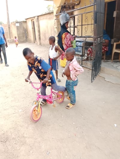 The scene is an unpaved outdoor area in the city of Masaka, Nigeria, illuminated by natural daylight. In the foreground, a boy, approximately 7-10 years old, wearing a dark blue patterned shirt and dark trousers with sandals, leans over a small pink bicycle with orange-rimmed wheels, hands on the handlebars, his left leg positioned over the frame, displaying a focused expression. To his immediate right stands a younger child, 3-5 years old, in an unbuttoned light pink shirt, blue jeans, and yellow slide sandals, observing the boy and bicycle.
Further back, a boy estimated 6-9 years old, in a white tank top and blue shorts, stands with hands on his hips. To the right, an adult female is positioned behind a dark metal gate with decorative scrollwork. She wears a black head covering, a multi-colored patterned dress, and a yellow skirt, with a dark plastic bag resting on her head. She faces towards the right or into a building. The background consists of light-colored, stucco-like residential buildings; one is yellow-tan with a dark doorway and window behind the gate, and another simpler structure is visible to the left. A light-colored plastic chair is near the doorway of the rightmost building. In the far left background, a partially visible individual in a blue shirt walks away, and a small child in a light-colored dress is seen walking in the distance. The ground is dry, light brown compacted dirt or sand.
