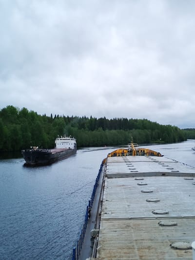 The image shows two barges navigating a canal in Depo, Russia. The sky is overcast. The foreground barge is light gray with rounded hatches on its surface and blue railings along its edge. The other barge is black and white, heading towards the camera in the distance. The canal is surrounded by dense green trees on both sides. The water is dark blue and rippled from the barges' movement.
