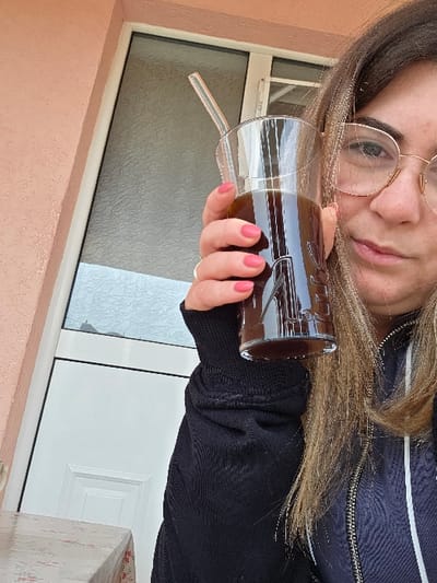The scene is set in Negorci, North Macedonia. A female subject is depicted from the chest up, looking directly at the camera. She has light brown hair and wears gold-rimmed spectacles. Her left hand, adorned with pink painted fingernails and a gold ring on the ring finger, holds a clear glass filled with a dark brown liquid. A transparent glass straw is inserted into the beverage. The subject wears a dark, long-sleeved garment, possibly a zippered jacket. The background features a textured, light peach-colored wall on the left, alongside a white-framed window with frosted glass. A white paneled door is partially visible below the window. A small section of a table with a floral-patterned tablecloth is visible in the lower-left foreground.