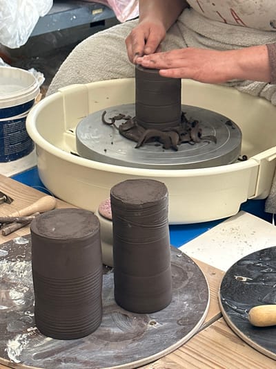 The scene depicts a pottery workshop in Mafra, Portugal, focusing on the creation of clay vessels. In the mid-ground, a person's hands, visibly coated with dark brown clay, are actively shaping a cylindrical piece of dark brown clay on a motorized pottery wheel. The clay cylinder exhibits distinct horizontal ridges, a result of the throwing process. Discarded scraps of dark brown clay surround the base of the active piece on the grey metal bat of the wheel. The wheel's splash pan is white plastic, situated above a blue surface.
In the foreground, to the left, two completed or partially completed cylindrical dark brown clay pieces stand upright on circular wooden bats. The leftmost cylinder displays prominent horizontal striations along its entire visible surface, while the adjacent cylinder also shows similar markings. To the left of these pieces, on a wooden surface, various pottery tools are laid out, including a wire cutting tool and other wooden implements. A white plastic bucket with blue labeling is partially visible in the upper left. A pink sponge is partially visible behind the foreground clay pieces. On the far right, another dark circular bat is visible, with a wooden tool handle resting upon it. The background is a blurry workshop environment.