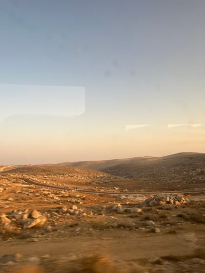 This wide-angle image depicts a barren, rocky, and hilly landscape near Carmel, Palestinian Territory, observed during what appears to be sunset or sunrise due to the warm, golden light illuminating the scene. The sky transitions from a pale blue at the top to a soft orange-yellow near the horizon. The photograph exhibits reflections in its upper half and slight motion blur in the immediate foreground, suggesting it was captured through a vehicle window.

The foreground and midground are characterized by dry, reddish-brown soil densely scattered with numerous light-colored boulders and sparse, low-lying, dry vegetation. A dark, paved road, likely asphalt, winds horizontally through the midground, extending from the lower left towards the right and disappearing behind a rise. Along this road, approximately in the center-left, two small, dark, indistinct figures are visible, suggestive of individuals walking or standing. The background consists of successive layers of rolling, arid hills, appearing progressively lighter and hazier with distance. On the far left horizon, faint structures, possibly buildings of a distant settlement, are discernible. No active interactions are apparent.