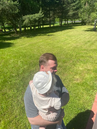 A bearded man, wearing a dark grey t-shirt and blue jeans, holds an infant or toddler outdoors in Копнино, Russia. The man's head is turned to his left, away from the direct camera view. The child, with their back to the camera, is wearing a light-colored bucket hat, a light-colored t-shirt, and striped shorts, resting against the man's chest. The environment consists of a vast, mown green lawn under direct sunlight, casting distinct shadows. A line of pine and deciduous trees forms the background, with a hammock visible hanging between trees on the left. A small section of a reddish-brown tiled or wooden structure is visible in the lower right foreground, partially obscured by a human shadow.