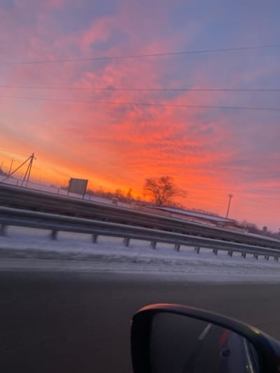 A vibrant, fiery sunset paints the sky in shades of orange and pink above a wintry roadside. Bare trees stand silhouetted against the horizon. A metal guardrail runs parallel to the road, edged with snow. Power lines stretch across the sky. The side mirror of a vehicle is visible in the foreground.