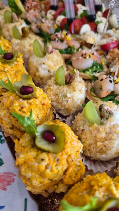 The image displays a close-up, top-down view of a platter of assorted appetizers. The appetizers are arranged in a visually appealing manner and include items such as small, round balls coated in nuts and seeds, topped with a grape slice and a pistachio. Other visible appetizers are topped with a slice of pickled cucumber and a single pomegranate seed, and a vibrant orange mixture that appears to be a shredded vegetable salad. In the background, there are also appetizers featuring shrimp, cherry tomatoes, and small mozzarella balls. The platter itself is set on a table covered with a white and pink patterned tablecloth. The overall presentation suggests a catering or party food spread. No specific location in Krauja, Latvia can be determined from the image.