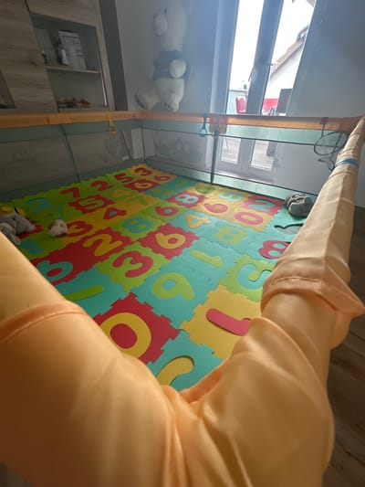 An indoor scene in Heinebach, Germany, shows a playpen with a numbered foam mat floor. The playpen has mesh sides with colorful attachments and a yellow frame. Scattered on the mat are two plush toys: one gray elephant and a smaller white and brown ball-shaped toy. A large teddy bear is visible in the background, mounted on a shelf. The room features a window or glass door that looks out onto a residential area with houses and a red chair on a balcony. The foreground is dominated by a large, draped orange fabric, likely part of the playpen structure.