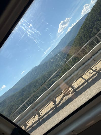 The image is a photograph taken from inside a vehicle, looking through a window. The view is angled, showing a mountainous landscape under a bright blue sky with scattered white clouds. In the foreground, a metal guardrail runs along the edge of what appears to be a road or bridge, casting shadows on the asphalt. The mountains are covered in dense green forest. The sun is shining brightly, causing lens flare and reflections on the window. The image appears to be taken on a sunny day in the vicinity of Vrachesh, Bulgaria.