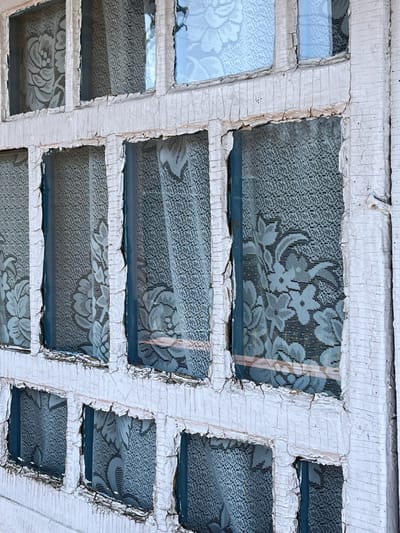 The image shows a grid of small, square windows with white, heavily cracked and peeling frames. The glass panes are partially obscured by lace curtains with a floral pattern. The environment is not directly visible beyond the curtains. The location is specified as Верхняя Уря, Russia, but there are no visual indicators in the image to confirm this.