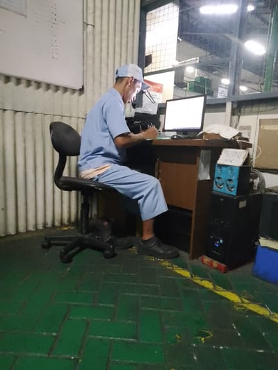 The image shows a person wearing a light blue uniform and a white and blue baseball cap, sitting at a desk in what appears to be a warehouse or factory setting. The person is looking down at a piece of paper and writing with a pen, with a computer monitor and keyboard in front of them. The desk is made of wood, and there is a black office chair.

To the right of the desk, there is a black computer tower with a blue speaker system on top. A small sign with handwritten text is placed on the speaker. The floor is green with yellow markings. In the background, a corrugated metal wall is visible, and a large window with metal bars looks out into another part of the facility, illuminated by bright artificial lights, suggesting it might be nighttime or an indoor environment with limited natural light. There's a whiteboard with some writing on the left side of the frame.

The overall scene suggests an individual working at a computer, possibly managing inventory or production data, in an industrial environment. The lighting indicates it could be late or an indoor work area.

Visible text: The sign on the speaker appears to read "SEDANG" and below it "RENC". The whiteboard on the left has handwritten notes, some of which look like numbers and letters. The computer monitor also displays text.