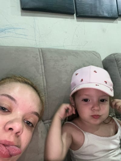 The image depicts two individuals, an adult and a young child, seated on a cushioned sofa. The adult, on the left, is closer to the camera and has blonde hair. The child, on the right, is wearing a pink cap adorned with small red fruit designs and a white tank top. The background shows a wall with some scuff marks, and above it, what appears to be a piece of dark artwork or a frame. The setting is indoors, likely a domestic environment. The image does not contain information about the city of Gorodets, Russia.