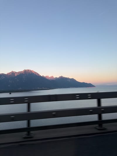 The image captures a panoramic view of the Alps and Lake Geneva, as seen from a bridge. The sky is a soft gradient of blue, transitioning to a pale orange at the horizon. A faint airplane contrail is visible in the upper right quadrant.
Dominating the middle ground are the majestic, snow-capped peaks of the Alps, bathed in the warm, rosy light of either sunrise or sunset. The lower slopes of the mountains are dark and shadowed, contrasting with the illuminated summits. A body of water, likely Lake Geneva, stretches across the lower half of the image, its surface calm and reflecting the light. Along the shoreline, a small town is nestled at the foot of the mountains. In the far right of the lake, a string of red lights can be seen, possibly marking buoys or a distant structure.
In the foreground, a dark metal guardrail runs horizontally across the frame, its bars and supports slightly blurred, suggesting movement as if captured from a moving vehicle. The road surface beneath the guardrail is also dark and shows motion blur.
The scene appears to be a scenic overlook, possibly part of a road or pathway constructed along the edge of Lake Geneva, offering a breathtaking vista of the natural landscape. The specific location within Villeneuve (VD), Switzerland, is suggested by the presence of the lake and the proximity to the Alps. The lighting indicates it is either early morning or late evening. There are no people or animals visible in the frame.
