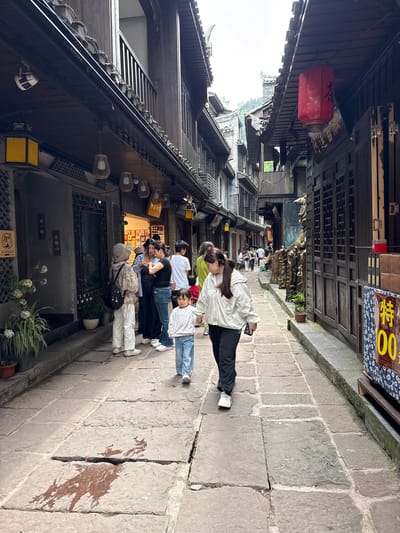 A photograph taken in Fenghuang, China, shows a stone pedestrian street lined with traditional wooden buildings. Several people are present, appearing to be tourists. They are positioned on the right side of the street. The street is paved with large, irregular stone slabs. Traditional Chinese architecture is visible, including overhanging eaves and decorative wooden features. A red lantern hangs from the building. A sign with yellow lettering hangs above the right side of the street. The environment is overcast, suggesting a cloudy day.