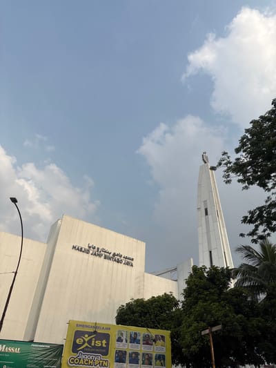 This is a low-angle, eye-level shot of the Masjid Jami' Bintaro Jaya in South Jakarta, Indonesia. The mosque's white facade is visible on the left, with Arabic script above the entrance reading "مسجد جامع بنتا رو جايا" and "MASJID JAMI' BINTARO JAYA" in English. A tall, slender white minaret with a patterned top stands to the right of the main building. Green trees and palm fronds partially obscure the mosque and minaret, and a street light is visible on the left. A yellow banner with the text "BIMBINGAN BELAJAR exist JAGONYA COACH PTN" and portraits of several individuals is displayed in the foreground, along with another green banner that reads "MASSAL" and other Indonesian text. The sky is blue with scattered white clouds.