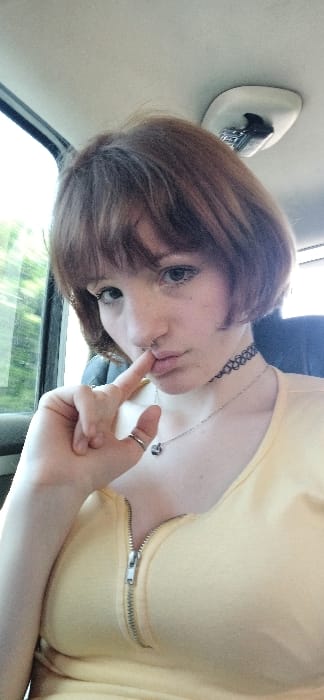 This image, captured inside a vehicle in Goričani, Montenegro, shows a female individual from the chest up. The subject has short, reddish-brown hair with bangs, a light complexion, and light-colored eyes. A small, circular ring is visible in a septum piercing. The left index finger is positioned against the upper lip, while the lips are slightly pursed. The individual wears a black, patterned choker necklace and a thin silver-colored chain with a dark pendant. A silver-colored ring is on the left ring finger. The subject is dressed in a yellow top featuring a central, partially unzipped silver-colored zipper. The vehicle's interior includes a light-colored headliner and an overhead light fixture on the upper right. Blurred green foliage is visible through the left side window.