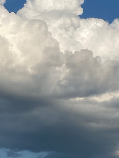 The image is a close-up, vertical shot of a sky filled with clouds. The clouds are varied in texture and color, with bright white, puffy formations in the upper portion contrasting with darker, grayer, and more ominous-looking clouds in the lower and middle sections. Some of the darker clouds appear dense and heavy, suggesting impending weather. A small patch of bright blue sky is visible at the very top left corner, peeking through the cloud cover. There are no people, animals, or man-made structures visible in the frame. The scene is purely atmospheric.