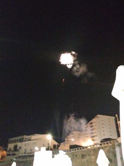 At night in Peso da Régua, Portugal, a firework explodes in the sky above a row of buildings. The burst is centered in the upper portion of the frame, producing a bright, diffused glow with visible smoke trailing behind it. Underneath the firework, a slender red trail marks its ascent. At the bottom of the image, multiple buildings are seen, partly obscured by what seems to be a wall or barrier in the foreground. The building to the right is taller, with several stories and windows visible, while the others are lower and spread out. Streetlights illuminate the buildings and create a hazy glow around them. The environment suggests an urban area during a celebratory event, as indicated by the fireworks display. The night sky occupies the majority of the frame, providing a stark contrast to the bursts of light.