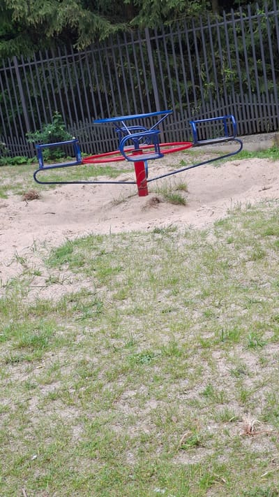 The image shows a red and blue merry-go-round on a playground. The merry-go-round is made of metal and has seats for children to sit on. It is located in Grójec, Poland, next to a black metal fence and some evergreen trees. The ground is a mixture of sand and grass. The scene appears to be outdoors during the daytime, with natural light illuminating the playground. There are no people visible in the image. The overall impression is one of a typical outdoor playground setting.