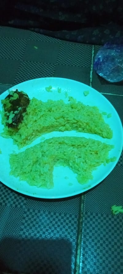 A plate of greenish rice and a dark, round item (possibly a meatball or vegetable fritter) rests on a checkered surface. A crumpled plastic bag is adjacent to the plate. A green object is to the bottom-right of the plate. The scene appears to be indoors, with a dark or shadowed background. The image location is Oshogbo, Nigeria.