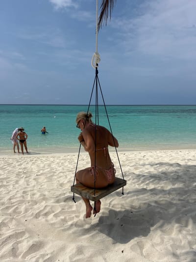 A woman wearing a bikini sits on a wooden swing at a beach. The swing is suspended by ropes from an unseen structure. The beach consists of white sand meeting a turquoise ocean. There are a few people visible in the background near the water. The sky is mostly clear with a few wispy clouds. The photo is taken from a slight angle, oriented vertically.