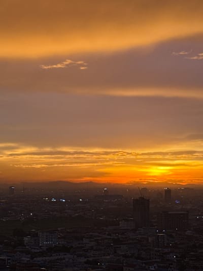 This media file depicts a panoramic sunset over a densely populated urban area. The sky is dominated by dramatic, layered clouds painted in vibrant hues of orange, yellow, and purple, indicative of a sunset. The light from the setting sun casts a warm glow on the clouds, creating a gradient from bright intensity at the horizon to softer tones further up.

Below the sky, a sprawling city is visible. The buildings are mostly dark and silhouetted, with scattered lights appearing as small, bright dots, suggesting that dusk is settling and artificial lighting is beginning to illuminate the urban landscape. Some buildings are more distinct, with visible windows and architectural details, including what appears to be a construction crane suggesting ongoing development. The dense arrangement of buildings implies a large city.

The perspective appears to be from an elevated viewpoint, possibly a tall building or a hill overlooking the city, allowing for a wide view of the skyline and the approaching twilight. The overall mood is one of awe and tranquility, with the natural beauty of the sunset contrasting with the ordered chaos of the urban environment. The time of day is clearly sunset, with the transition from daylight to night in progress. The weather appears clear enough for a dramatic sunset. The location is identified as Khan Mean Chey, Cambodia, which is a district within Phnom Penh, the capital city. No text is visible in the media file.