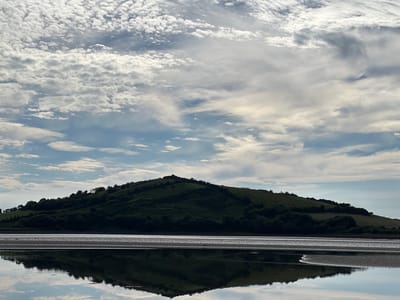 This is a wide shot of a landscape scene in Dunuisce, Ireland. The image is divided into three horizontal sections: the sky, the land, and the water. The sky is filled with a variety of clouds, from small, fluffy cumulus clouds to larger, wispy cirrus clouds. The land is a verdant green hill covered in trees, sloping down to the water. The water is calm and reflects the sky and the hill, creating a mirror image of the landscape. The overall impression is one of peace and tranquility.