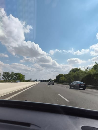 The image depicts a view from inside a vehicle traveling on a highway. The sky is blue with scattered white clouds. On the right side of the highway, there is a dense line of green trees. In the distance, several wind turbines are visible. On the highway, several cars are driving in the same direction. The foreground shows the dashboard of the vehicle. The image was taken in Châtenay, France.
