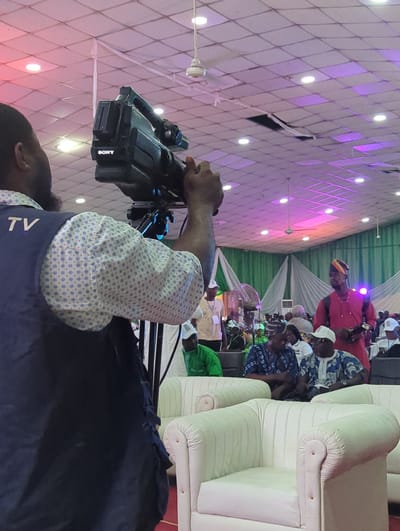 The image depicts a scene in Ungwan Liman, Nigeria, showing a cameraman operating a Sony video camera on a tripod. He is wearing a blue shirt with "TV" printed on it, as well as a patterned shirt underneath. The camera is focused on a group of people seated in the background, including one man wearing a red robe and hat. The setting appears to be an event or gathering, as indicated by the seating arrangement with white cushioned chairs and the presence of a decorated backdrop featuring green and white fabric. The ceiling is white with a diamond pattern and multiple recessed lights that cast a pinkish hue.